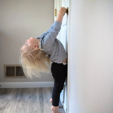Child playing with door knob, showing curiosity while hanging from door, with sandy blonde hair and casual gray outfit.