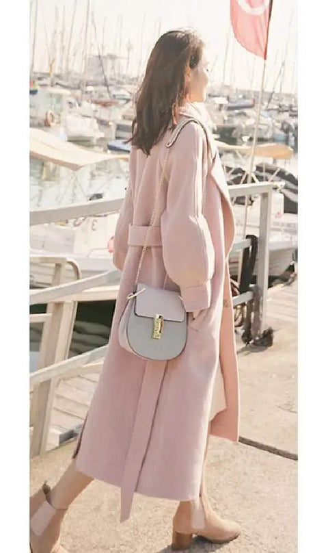 Woman wearing a long pink coat and carrying a gray handbag, walking by boats at a marina.