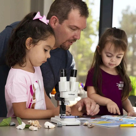 Portable Binocular Stereo Microscope With LED Illumination And 10x Widefield Eyepieces Built For Hands On Learning At Home Or In The Classroom Perfect For Checking Biological Specimens Rocks Stamps Jewelry And Other Tiny Objects. White binocular microscope with LED light used by children at a table.