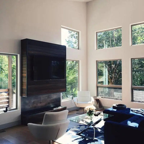 Modern living room with large windows, featuring a wood accent wall, TV, white chairs, and glass coffee table.
