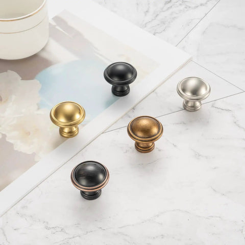 Five different finishes of cabinet knobs laid on a marble surface, including Ten Pack One And One Quarter Inch Antique Brass Kitchen Cabinet Knobs.