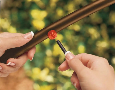 Hands installing 2 GPH Pressure Compensating Drip Emitter for Spot Watering, self-piercing design into tubing with greenery background