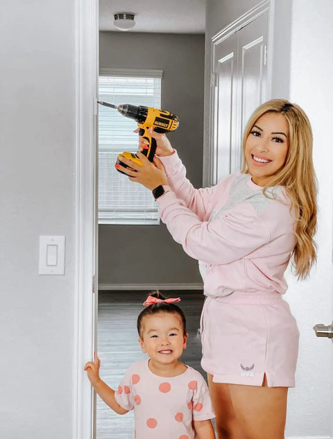 Woman holding a drill near a door, child smiling beside her in pink outfit. Home improvement scene.