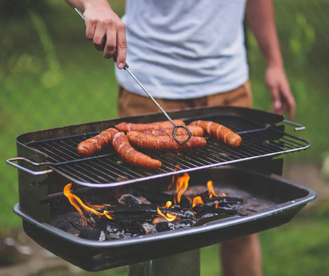 Man grilling sausages over open fire on an outdoor barbecue grill in a backyard setting.