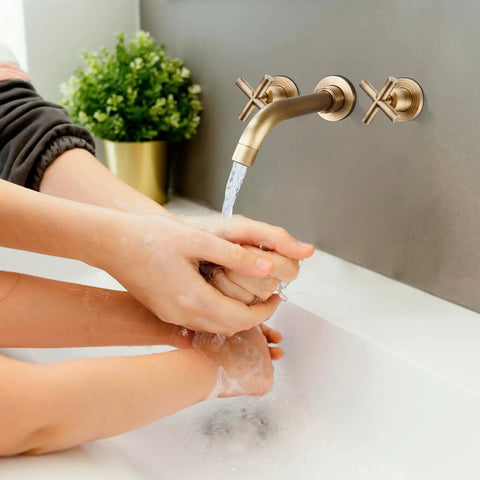 Hands being washed under wall mounted bathroom sink faucet with dual cross handles, 3 hole widespread mixer tap in brass finish.