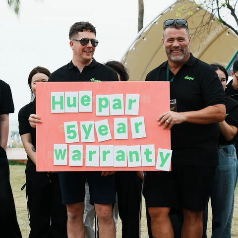 Two men holding a sign that says 'Hue par 5 year warranty' in a group setting at an outdoor event.