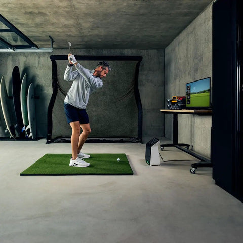 Man using Premium Golf Launch Monitor And Simulator Bundle With A 10 Inch Color Touchscreen, Over 15 Metrics, GPS Virtual Course Overview in garage setup.