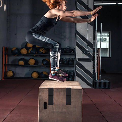 Person performing box jump exercise in gym with wall-mounted medicine balls and kettlebells in the background.