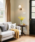 Living room with beige sofa, black door, floral cushions, wooden table, vase with flowers, and visible light switch wall plate.
