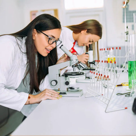 Two women in lab coats examining samples with a microscope and a Durable Metal Lab Beaker Drying Holder And Test Tube Rack Draining Stand For Glassware Organization 11x11 Inch.
