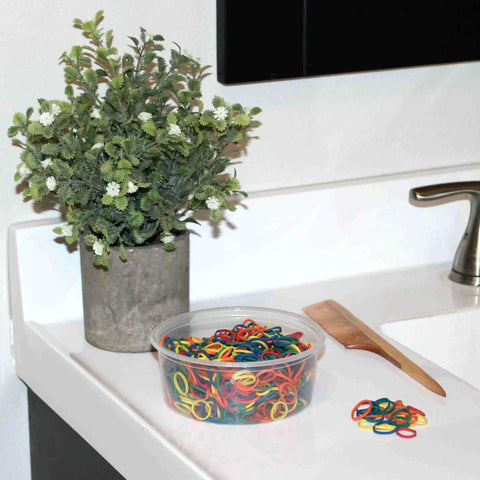 Dope Half Inch Rainbow Mix Small Rubber Bands For Hair Pack 500 Count in a clear tub next to a plant and a comb on a bathroom counter.