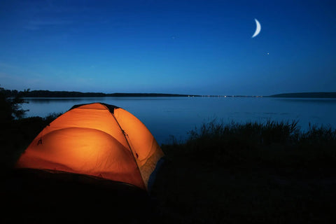 A glowing tent by a lakeside at night under a crescent moon, illustrating the Compact LED Flashlight With 1-Cell AAA Battery use in camping.