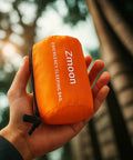 Hand holding a pack of Emergency Mylar Sleeping Bag Set For Camping Waterproof Thermal Bivy Sacks Bivvy Bags in bright orange.