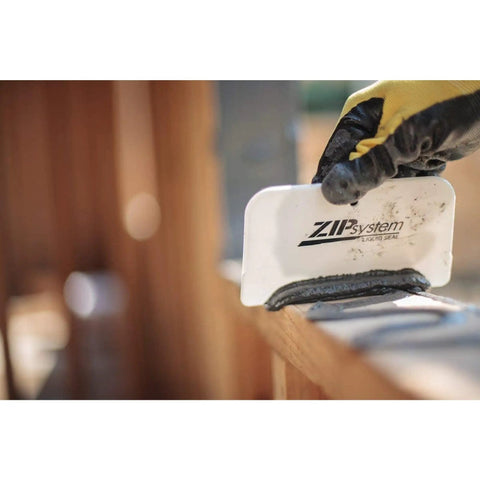 Person applying Fluid Applied STPE Liquid Flashing Sealant In 29 Oz Cartridge with a scraper on a wooden surface.