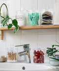 Super Practical Four Pack Clear Acrylic Qtip Holder Dispensers With Lids For Organizing Cotton Swabs Balls Pads And Floss Picks Comes With Labels In Two Sizes Twelve Ounce And Ten Ounce on a bathroom shelf.