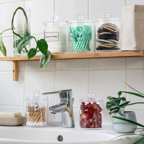 Super Practical Four Pack Clear Acrylic Qtip Holder Dispensers With Lids For Organizing Cotton Swabs Balls Pads And Floss Picks Comes With Labels In Two Sizes Twelve Ounce And Ten Ounce on a bathroom shelf.