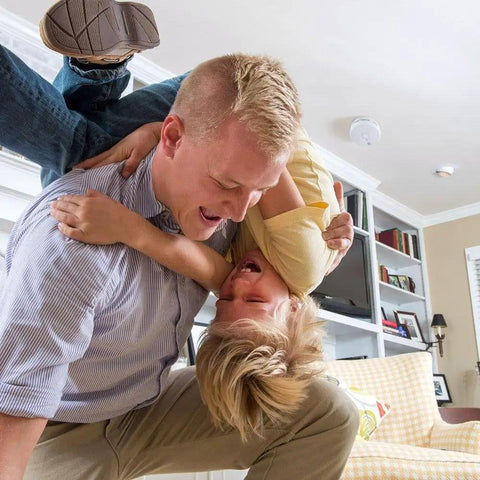 Father playing joyfully with child in living room featuring Set It And Forget It With This Totally Worry Free 10 Year Sealed Lithium Photoelectric Smoke Detector.