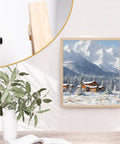 Wall-mounted painting of snowy mountain landscape with evergreen trees, framed in light wood; partial view of a black hook.