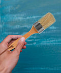 Hand holding 2 Piece Wood Handle Paint Stain Brushes With Oval Synthetic Bristles For Wood Stain And Wall Paint Deck Brush For Furniture And Wall Treatments This Pro Painting Tools Set Delivers Smooth Finishes And Easy Cleanup in front of blue painted wall