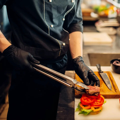 Front view: Schneider black vinyl exam gloves worn by a chef, handling food with tongs over a burger.