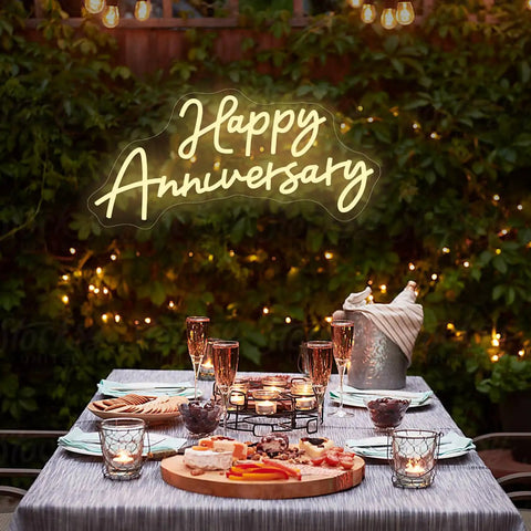 Happy Anniversary neon sign glowing over a romantic outdoor table setting with candles and drinks.
