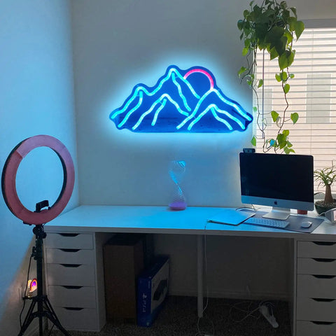 Blue neon mountain wall decor above a desk with computer, ring light, and potted plant in a modern workspace.