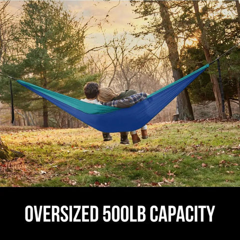 Durable Camping Hammock Built Tough With Heavy Duty Tree Straps Holds Up To 500 Lbs Steel Carabiners For A Secure Hang Easy Setup For Outdoor Trips Hiking And Backpacking Double Size Portable Blue, hanging between trees outdoors, with two people reclining.