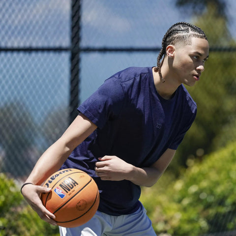 A person playing basketball outdoors with the Official DRV Series Basketball 27.5 Inch Size 5 In Brown With Rugged Outdoor Cover And NBA Branding.