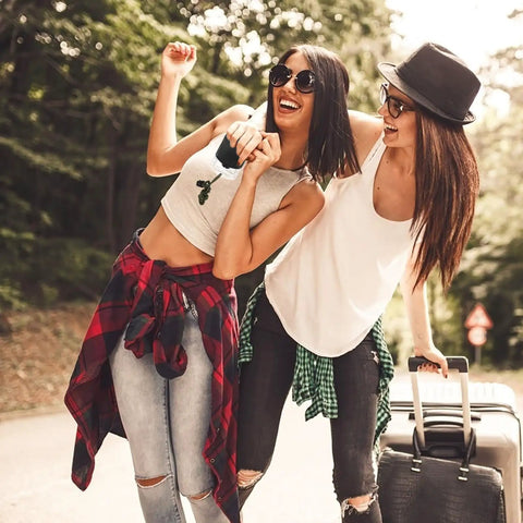 Two people smiling on a wooded path, casual summer attire, carrying a suitcase.