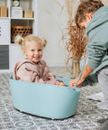 Child inside a blue plastic bin being pulled by another child, indoors on patterned flooring.