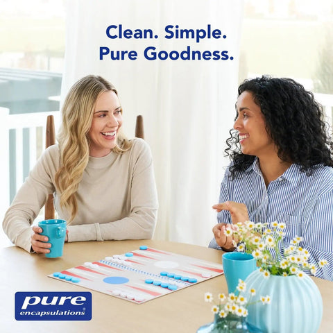 Two women sitting at a table with board game, blue mugs, and branded Pure Encapsulations logo, floral vase in foreground.