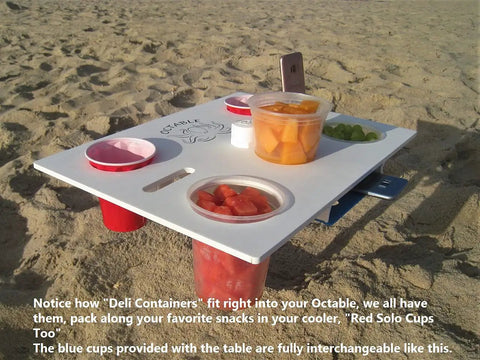 Portable Beach Table With Two Phone Slots, Lightweight Yet Sturdy And Durable, Must Have For Beach, Boat, Pool, Backyard, RV And Camping, Keeps Drinks And Phones Off The Sun With Built In Tray For Stash And Gear on sand with deli containers and blue cups.