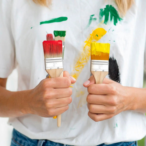 Person holding two paint brushes with red and yellow tips, showing the Ergonomic Thick Wood Handle Flat Paint Brush Set Of 30 Pieces