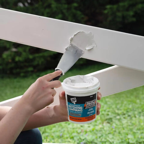 Applying Weatherproof Patching Compound 16oz White For Drywall And Masonry Repairs to an outdoor wooden surface with a putty knife.