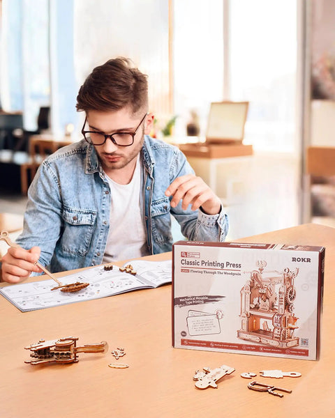 3D Puzzle Kit For Adults Wooden Printing Press Model Building Kit With 303 Pieces, DIY Wood Craft, STEM Hobby Toy, Includes Illustrated Manual, Screwdriver, Spring, And Custom Rubber Stamp Set For Creative Hands On Fun. Man assembles wooden printing press kit with instructions