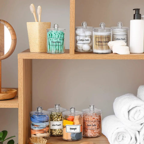 Super Practical Four Pack Clear Acrylic Qtip Holder Dispensers With Lids For Organizing Cotton Swabs Balls Pads And Floss Picks Comes With Labels In Two Sizes Twelve Ounce And Ten Ounce on wooden shelf.