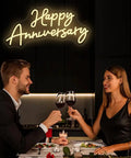 Couple toasting with glasses under 'Happy Anniversary' neon sign in dimly lit restaurant setting.