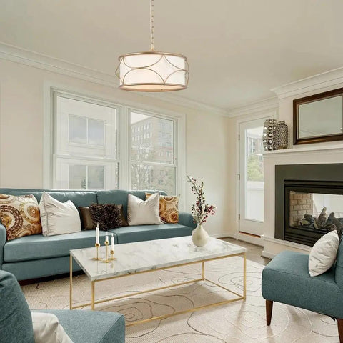 Gold Drum Pendant Chandelier Light Fixture with White Fabric Shade hanging in a modern living room above a marble coffee table.