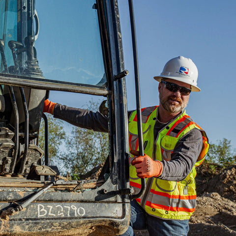 A man in a SV55 Class 2 Heavy Woven Two Tone Engineer Vest With Padded Neck For Extra Weight In Cargo Pockets ANSI ISEA 107 Class 2 Hi Vis Worksite Ready Rugged Durable Construction Green Large, standing by a construction vehicle on a worksite.