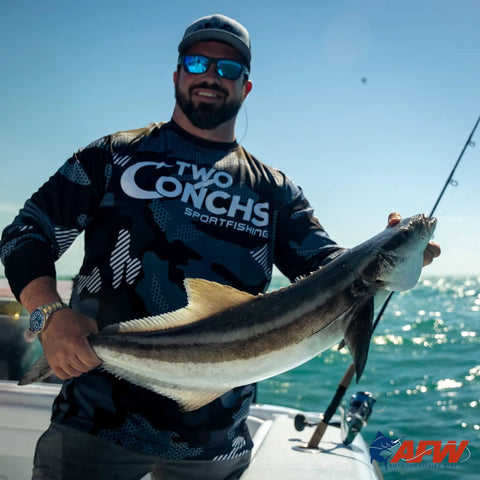 Person holding a large fish on a boat with a fishing rod, wearing Two Conchs Sportfishing shirt under a clear blue sky.