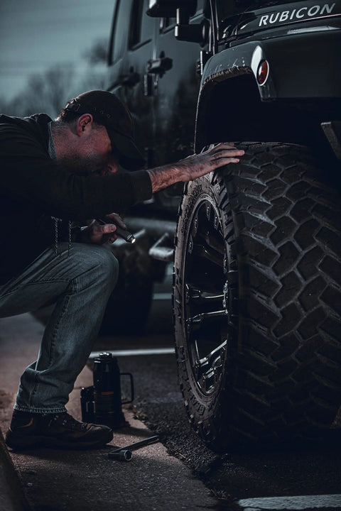 Man inspecting large tire of a parked off-road vehicle at dusk.