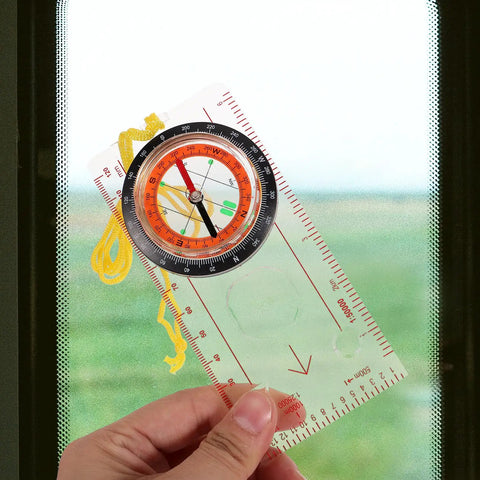 Hand holding Pocket Multitool Hiking Compass And Measuring Ruler For Outdoor Navigation against a window