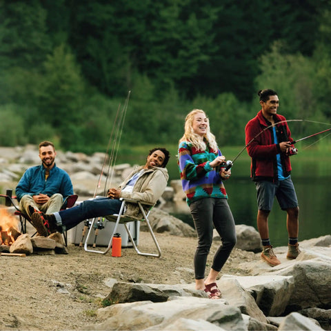 Group of people fishing by a lakeside with rocks and trees in the background, relaxed and enjoying a peaceful outdoor setting.