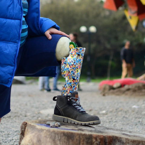A child with a colorful patterned prosthetic leg resting on a log, wearing a black boot, outdoors.