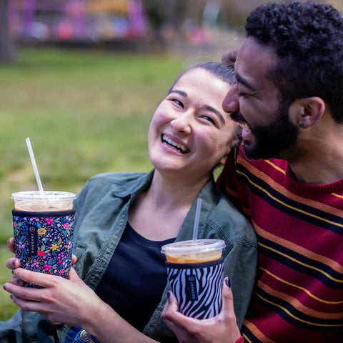 Couple enjoying iced coffee in Premium Neoprene Iced Coffee Sleeve, holds 22 to 28 oz cups, outdoor setting.