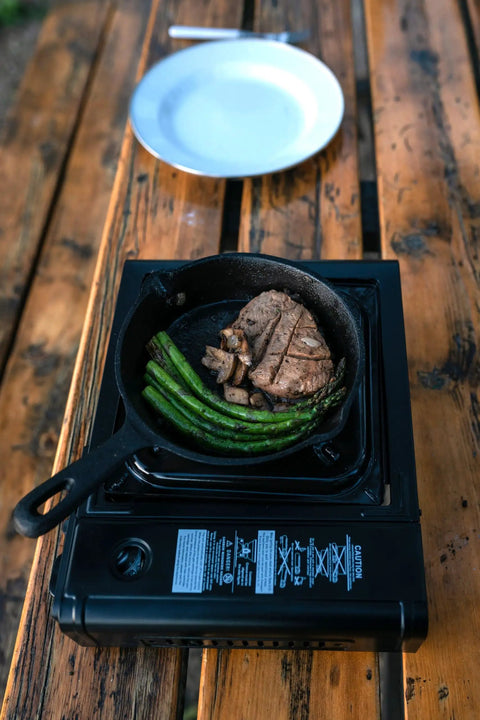 Portable Outdoor Butane Stove For Camping And Catering On The Go With Electronic Ignition Ultra Quiet One Burner Cooker In Black Includes Hard Case Works With 8 Oz Butane Canisters For BBQs showing steak and asparagus being cooked.