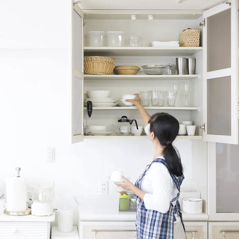 White kitchen wall cabinet with open doors and shelves; glassware, bowls and baskets, while a woman cleans.