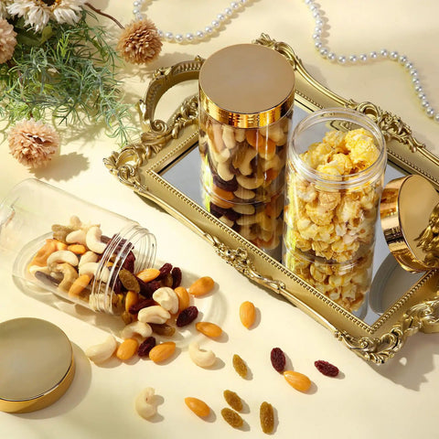 Hella Durable 12 Pack Of 8 Oz Round Clear Plastic Jars With Secure Twist Lids, Premium Pet Containers For Slime, Cosmetics, Travel Kit, And Pantry Storage With Gold Caps filled with nuts and popcorn on a mirror tray.