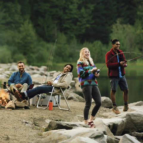 Group of friends fishing by a lake with trees in the background, some sitting near a campfire, others standing with fishing rods.