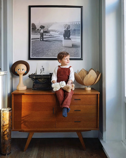 Child sitting on vintage cabinet with typewriter, hat mannequin, and decorative lamp in a cozy room.